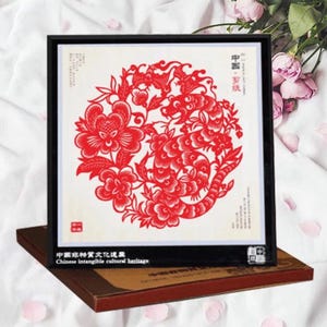 May include: A framed red paper cut-out art piece featuring a floral and dragon design. The artwork is set against a white background and framed in black. The text "Chinese intangible cultural heritage" is visible.
