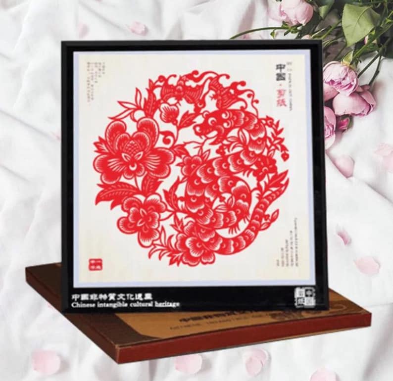 May include: A framed red paper cut-out art piece featuring a floral and dragon design. The artwork is set against a white background and framed in black. The text "Chinese intangible cultural heritage" is visible.