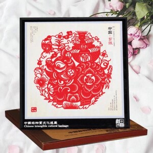May include: A framed red paper cut-out art piece featuring a pig and floral design. The artwork is set against a white background and framed in black. The text "Chinese intangible cultural heritage" is visible.