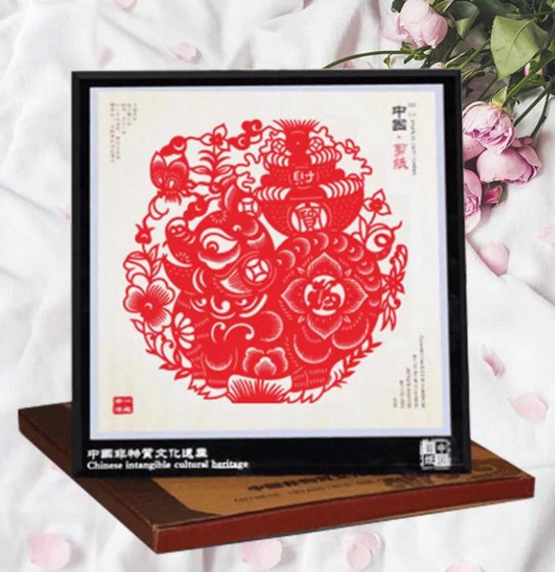 May include: A framed red paper cut-out art piece featuring a pig and floral design. The artwork is set against a white background and framed in black. The text "Chinese intangible cultural heritage" is visible.