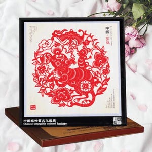 May include: Framed red paper cut art featuring a detailed design of a rat surrounded by floral and cloud motifs. The artwork is set against a cream background and framed in black. The frame rests on a brown box.