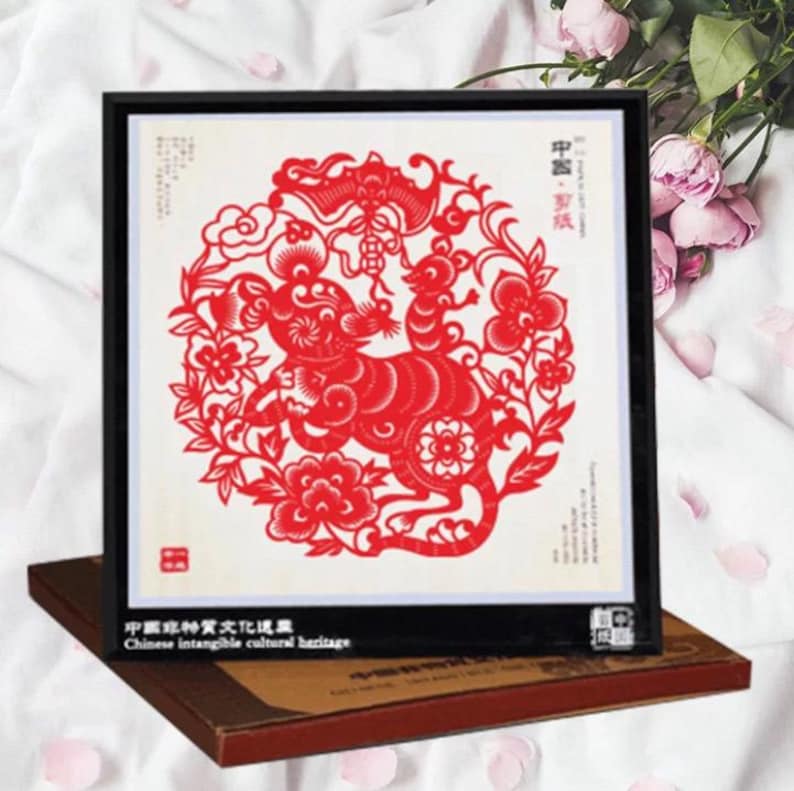 May include: Framed red paper cut art featuring a detailed design of a rat surrounded by floral and cloud motifs. The artwork is set against a cream background and framed in black. The frame rests on a brown box.