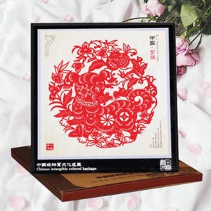 May include: A framed red paper cut-out art piece featuring a detailed design of a mythical creature and floral elements. The artwork is set against a white background and framed in black. The text "Chinese intangible cultural heritage" is visible.