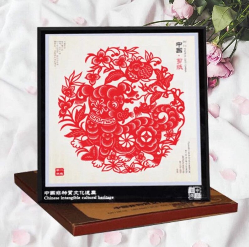 May include: A framed red paper cut-out art piece featuring a detailed design of a mythical creature and floral elements. The artwork is set against a white background and framed in black. The text "Chinese intangible cultural heritage" is visible.