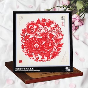 May include: A framed red paper cut-out of a ram, surrounded by floral and bird designs. The artwork is displayed on a brown box, with text in Chinese and English. The frame is black.
