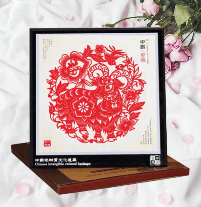 May include: A framed red paper cut-out of a ram, surrounded by floral and bird designs. The artwork is displayed on a brown box, with text in Chinese and English. The frame is black.