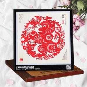 May include: A framed red paper cut-out with a circular design featuring a stylized animal, flowers, and leaves. The frame is black, and the artwork is displayed on a brown base. Text in Chinese and English is visible.