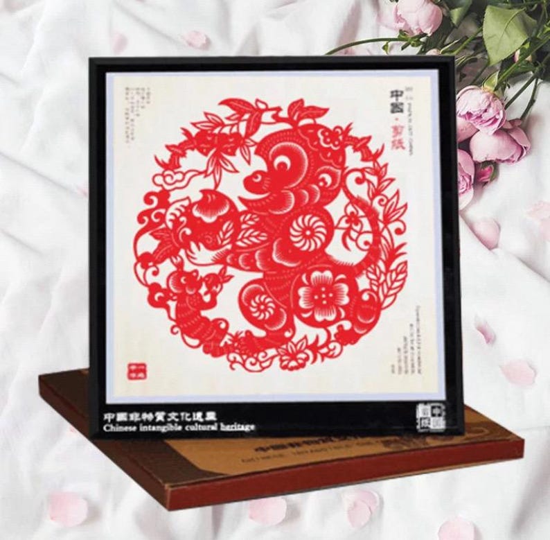 May include: A framed red paper cut-out with a circular design featuring a stylized animal, flowers, and leaves. The frame is black, and the artwork is displayed on a brown base. Text in Chinese and English is visible.