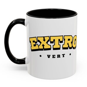 Może przedstawiać: Biały ceramiczny kubek z czarnym uchwytem i wnętrzem. Kubek zawiera słowo "EXTRO" w żółtych blokowych literach z czarnym obrysem oraz słowo "VERT" w kolorze czarnym poniżej. Stylowy kubek do kawy do codziennego użytku.
