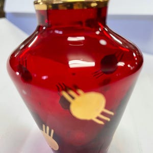 May include: A red glass vase with a gold rim and gold abstract designs. The vase has a bulbous shape and a narrow neck. The gold designs are scattered around the vase. Likely vintage.