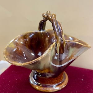 Puede incluir: Una cesta de cerámica marrón con asa. La cesta tiene un borde ancho y acampanado y una base de pedestal. El asa es un diseño decorativo en bucle. El esmalte tiene un acabado brillante con variaciones de marrón y beige.