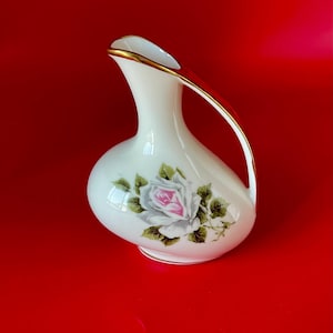 May include: A white ceramic pitcher with a gold rim and handle. The pitcher features a hand-painted pink and white rose with green leaves. The background is a solid red.