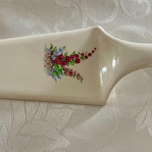 Op de afbeelding: Crèmekleurige keramische taartschep met een bloemenontwerp met rode, blauwe en groene bloemen. De handgreep is lichtgeel. Een decoratief serveerstuk voor taarten en cakes.