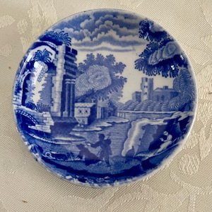 May include: A small, round blue and white ceramic dish with a detailed landscape scene. The design features buildings, trees, and figures, all in a classic blue and white colour scheme. The dish has a slightly raised edge.