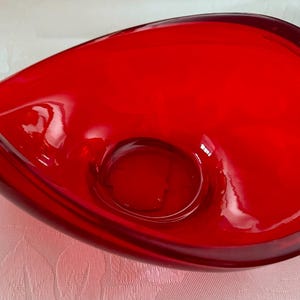 May include: A striking red glass bowl with an unusual, flowing form. The bowl has a central, circular recess and a smooth, shiny surface. The design is reminiscent of a stylized leaf or petal.