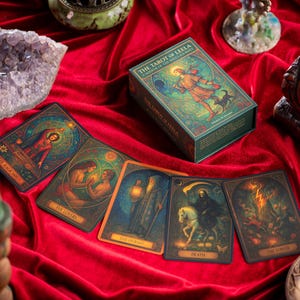 May include: A collection of tarot cards and a box set on a red velvet surface. The box is green with the title "The Tarot of Leela". Several cards are displayed, each with unique illustrations and titles. A wooden Buddha statue and amethyst geode are also present.