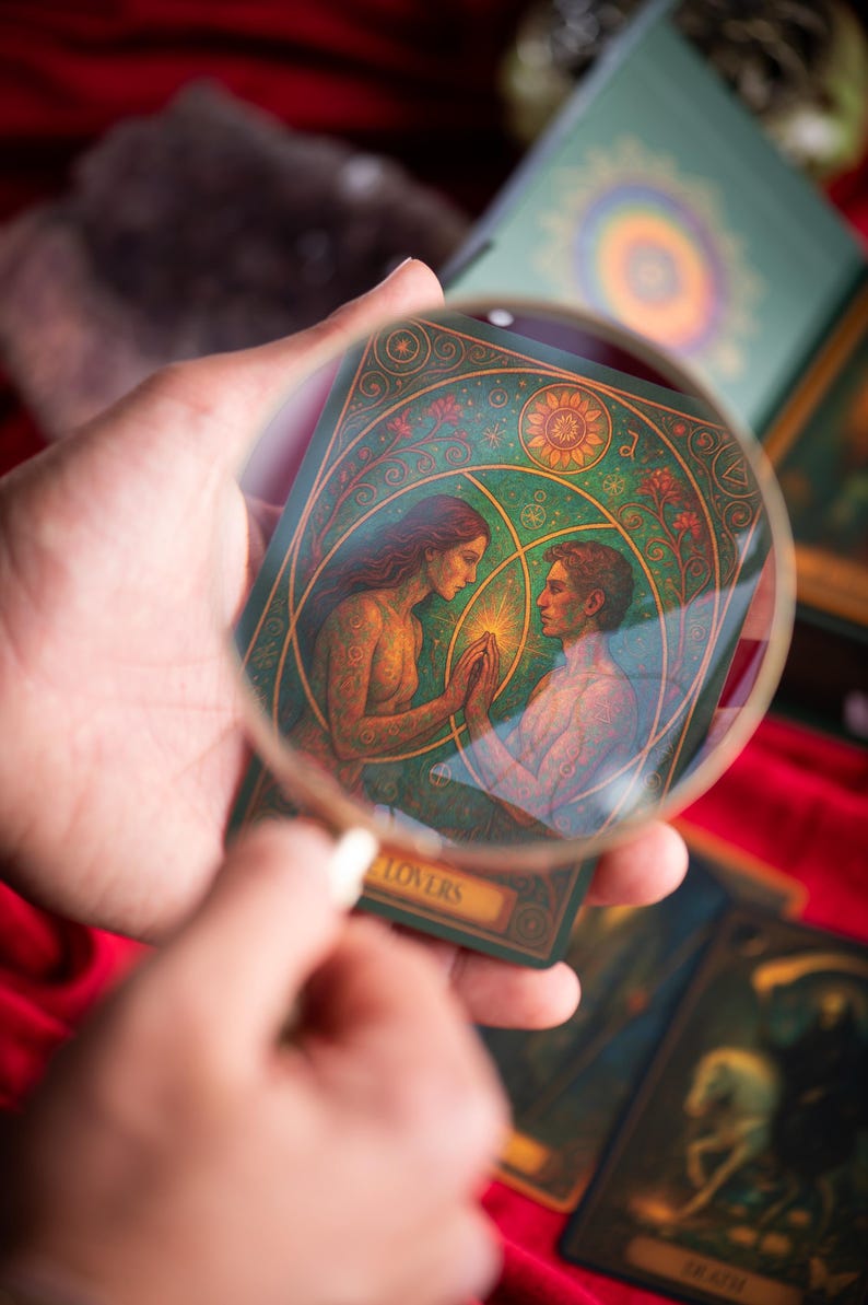 Pu&ograve; includere: Una carta dei tarocchi raffigurante due figure che si tengono per mano, vista attraverso una lente d'ingrandimento. La carta ha uno sfondo verde con dettagli dorati e la parola "LOVERS" in basso. Altre carte dei tarocchi e una scatola sono sullo sfondo.