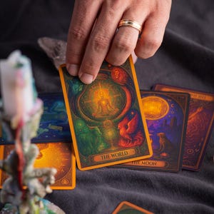 Pu&ograve; includere: Una mano che tiene una carta dei tarocchi con la scritta "THE WORLD" con un'illustrazione dettagliata di una figura in meditazione. Sono visibili altre carte con immagini celesti e simboliche, che suggeriscono un tema di divinazione.