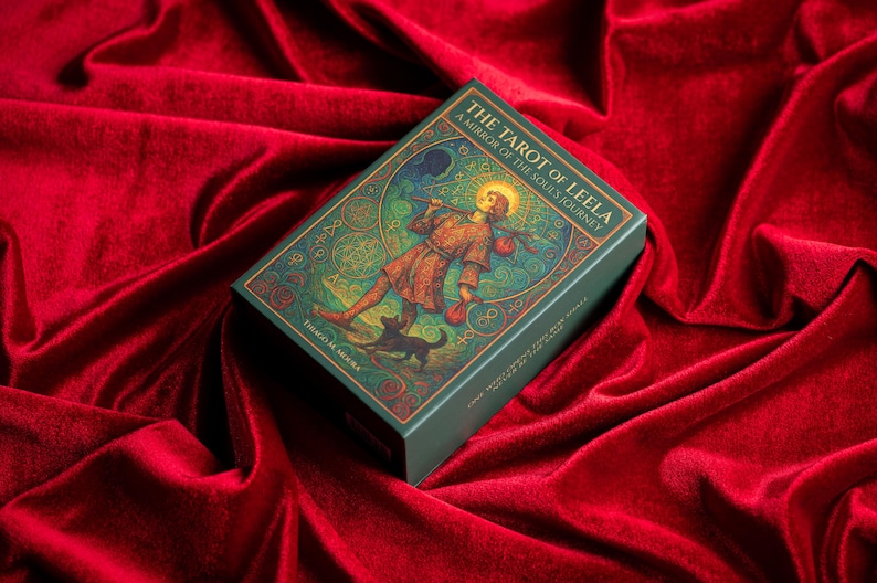 Pu&ograve; includere: Un mazzo di carte dei tarocchi intitolato "The Tarot of Leela" con una scatola verde con un'illustrazione di una figura con un alone, un cane e del testo. Il mazzo poggia su una superficie di velluto rosso.