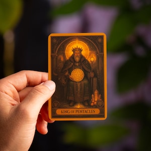 Pu&ograve; includere: Una carta dei tarocchi raffigurante il Re di Denari. La carta ha un bordo arancione e raffigura un re con una corona, che tiene un pentacolo d'oro. La carta &egrave; tenuta in mano su uno sfondo sfocato.