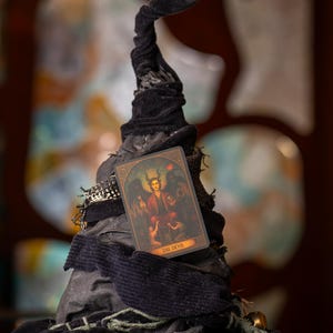 Pu&ograve; includere: Un cappello da strega nero con la parte superiore attorcigliata, adornato con tessuto scuro ed elementi decorativi. Una carta dei tarocchi raffigurante una figura di diavolo &egrave; posta sul cappello. La carta recita "THE DEVIL".
