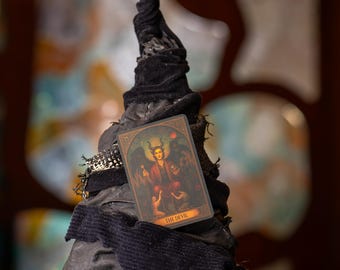 Può includere: Un cappello a punta in tessuto nero con una consistenza usurata e stratificata, caratterizzato da una carta dei tarocchi intitolata The Devil fissata sul davanti. Il cappello ha una corona alta leggermente curva e una tesa larga e arricciata.