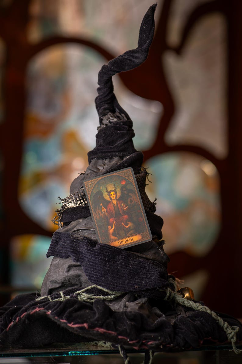 Pu&ograve; includere: Un cappello da strega nero con la parte superiore attorcigliata, adornato con tessuto scuro ed elementi decorativi. Una carta dei tarocchi raffigurante una figura di diavolo &egrave; posta sul cappello. La carta recita "THE DEVIL".