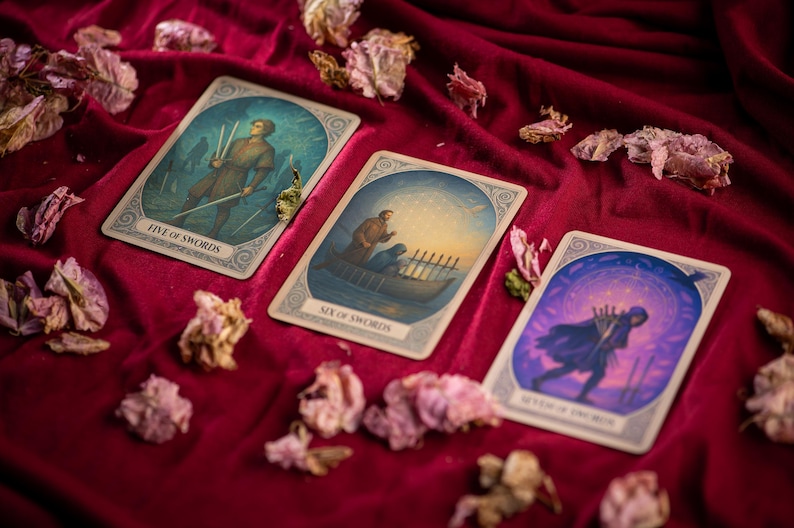 Pu&ograve; includere: Tre carte dei tarocchi con illustrazioni e testo su una superficie di velluto rosso, circondate da fiori secchi. Le carte sono etichettate come "Five of Swords", "Six of Swords" e "Seven of Swords", ognuna raffigurante figure con spade.