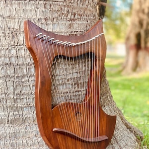 May include: A dark brown wooden lyre, a stringed instrument, leaning against a tree trunk. The lyre has a curved shape, with numerous strings stretched across its body. The instrument's design includes a sound hole and decorative pegs.