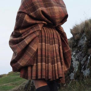 Puede incluir: Un kilt a cuadros marrón y rojo, con un chal a juego. El kilt está plisado y se lleva con calcetines altos grises oscuros y zapatos de cuero marrón. Adecuado para la vestimenta tradicional escocesa.