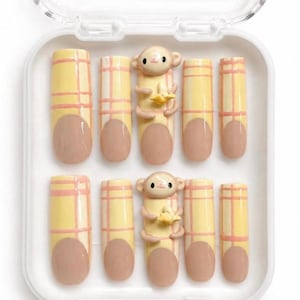 May include: A set of ten press-on nails in a white plastic storage case. The nails feature a yellow and pink plaid pattern with nude-coloured tips. Two of the nails are adorned with a three-dimensional sculpted monkey figure holding a yellow banana.