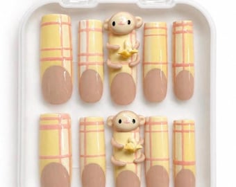 Puede incluir: Un juego de diez uñas postizas en un estuche de plástico blanco. Las uñas presentan un patrón de cuadros amarillos y rosas con puntas de color carne. Dos de las uñas están adornadas con una figura de mono en relieve que sostiene un plátano.