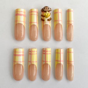 Könnte beinhalten: Set mit zehn künstlichen Nägeln. Jeder Nagel hat eine hellbraune Basis mit einem gelb-rosa Karomuster. Ein Nagel hat eine kleine Affenfigur, die eine Banane hält. Die Nägel sind oval geformt.