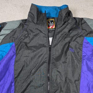 May include: A vintage windbreaker jacket in black, blue, and purple with a full zip. The jacket displays the USA Olympic logo and text on the label and a small logo on the chest. It has a high collar and a colour-blocked design.