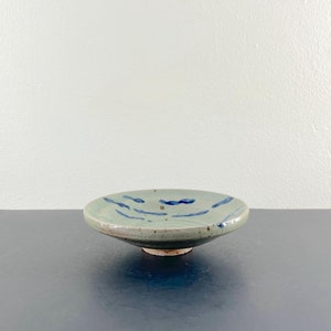 Pode incluir: Uma pequena tigela de cerâmica azul claro com uma borda ligeiramente elevada e um esmalte azul e branco. A tigela tem um design simples pintado à mão de linhas azuis.