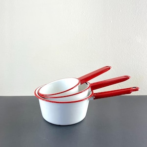 May include: Three white enamel saucepans with red rims and handles. The saucepans are nested together.