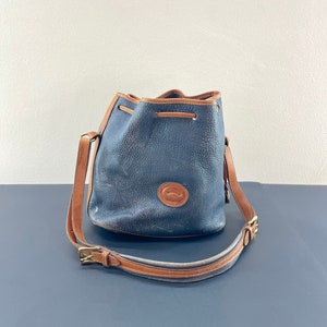 May include: A blue leather bucket bag with a brown leather strap and a brass buckle. The bag has a drawstring closure and a logo on the front.