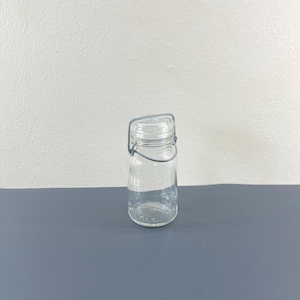 Puede incluir: Un tarro de vidrio transparente con un asa de alambre. El tarro está vacío y tiene una superficie texturizada.