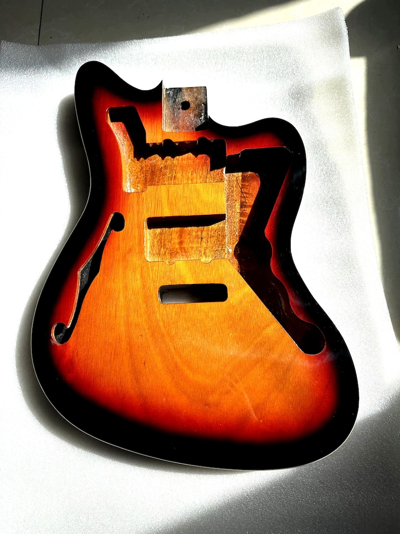 May include: A hollow-body electric guitar body with a sunburst finish. The body is made of wood and features a black edge, orange and red gradient, and a natural wood grain center. It has cutouts for the pickups, bridge, and f-holes.