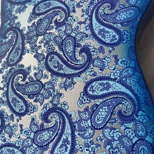 K&ouml;nnte beinhalten: Nahaufnahme eines blauen Paisley-Musters auf einer Oberfl&auml;che. Das Design zeigt gro&szlig;e, tropfenf&ouml;rmige Paisley-Motive in verschiedenen Blaut&ouml;nen, vor einem silbernen Hintergrund. Das Muster ist detailliert und komplex, mit einer Mischung aus floralen und abstrakten Elementen.