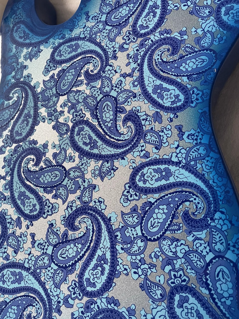 K&ouml;nnte beinhalten: Nahaufnahme eines blauen Paisley-Musters auf einer Oberfl&auml;che. Das Design zeigt gro&szlig;e, tropfenf&ouml;rmige Paisley-Motive in verschiedenen Blaut&ouml;nen, vor einem silbernen Hintergrund. Das Muster ist detailliert und komplex, mit einer Mischung aus floralen und abstrakten Elementen.