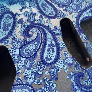 K&ouml;nnte beinhalten: Nahaufnahme eines Gitarrenkorpus mit blauem Paisley-Muster. Das Design zeigt gro&szlig;e, wirbelnde Paisley-Formen in Blau- und Wei&szlig;t&ouml;nen vor einem silbernen Hintergrund. Der Gitarrenkorpus hat schwarze Aussparungen.
