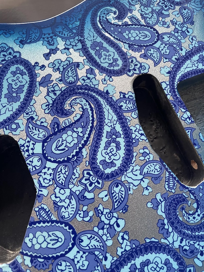 K&ouml;nnte beinhalten: Nahaufnahme eines Gitarrenkorpus mit blauem Paisley-Muster. Das Design zeigt gro&szlig;e, wirbelnde Paisley-Formen in Blau- und Wei&szlig;t&ouml;nen vor einem silbernen Hintergrund. Der Gitarrenkorpus hat schwarze Aussparungen.