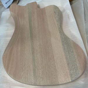 May include: Unfinished wooden guitar body with a natural wood grain pattern. The body is shaped with curves and a cutaway for the neck. The wood has a light brown color with streaks of gray and pink.