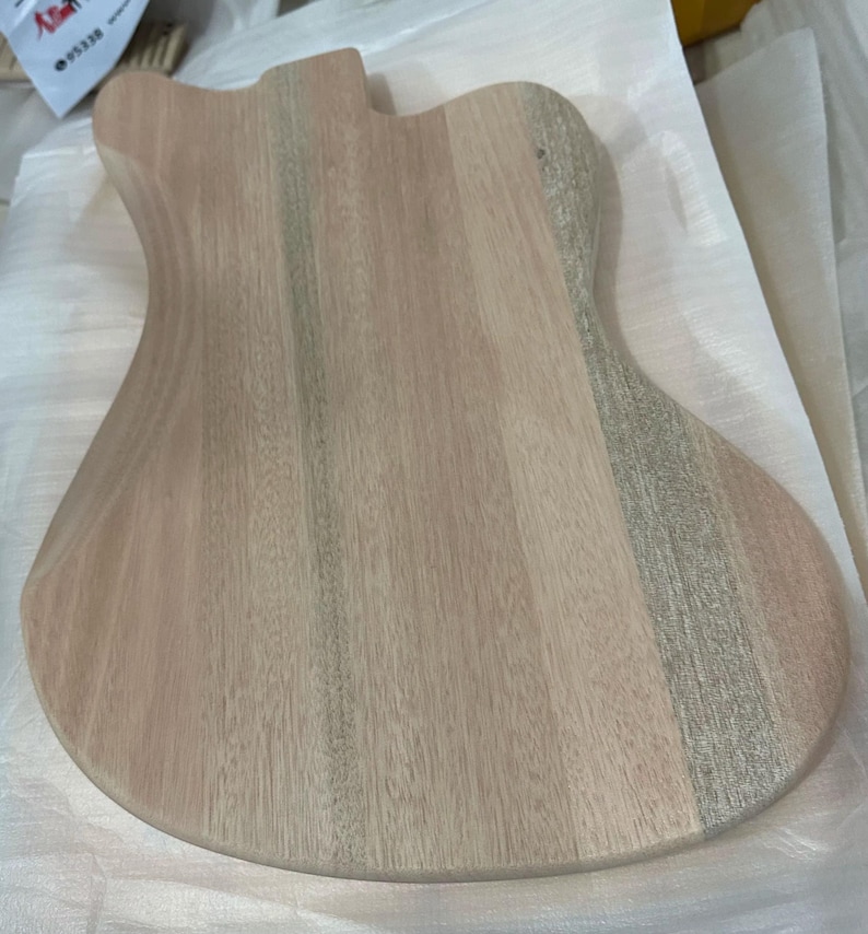 May include: Unfinished wooden guitar body with a natural wood grain pattern. The body is shaped with curves and a cutaway for the neck. The wood has a light brown color with streaks of gray and pink.