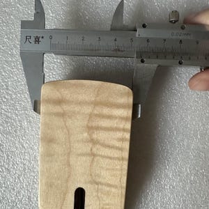 以下が含まれることがあります： 明るい色の木製ギターネックを、銀色のノギスで測定しています。ノギスは約5cmの測定値を示しています。ネックには、暗い長方形のスロットと自然な木目があります。