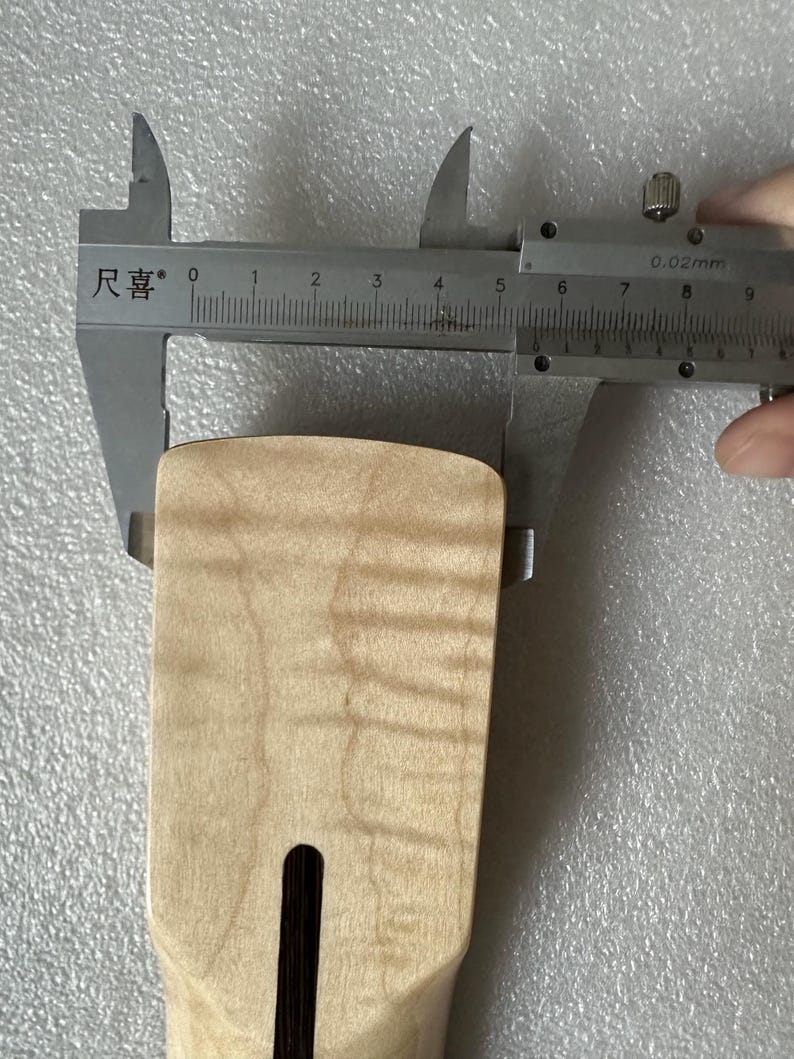 以下が含まれることがあります： 明るい色の木製ギターネックを、銀色のノギスで測定しています。ノギスは約5cmの測定値を示しています。ネックには、暗い長方形のスロットと自然な木目があります。