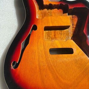 May include: A hollow-body electric guitar body with a sunburst finish. The body is unfinished, revealing the wood grain. The color transitions from black to red to orange and yellow. The guitar has a classic f-hole design and cutaways.