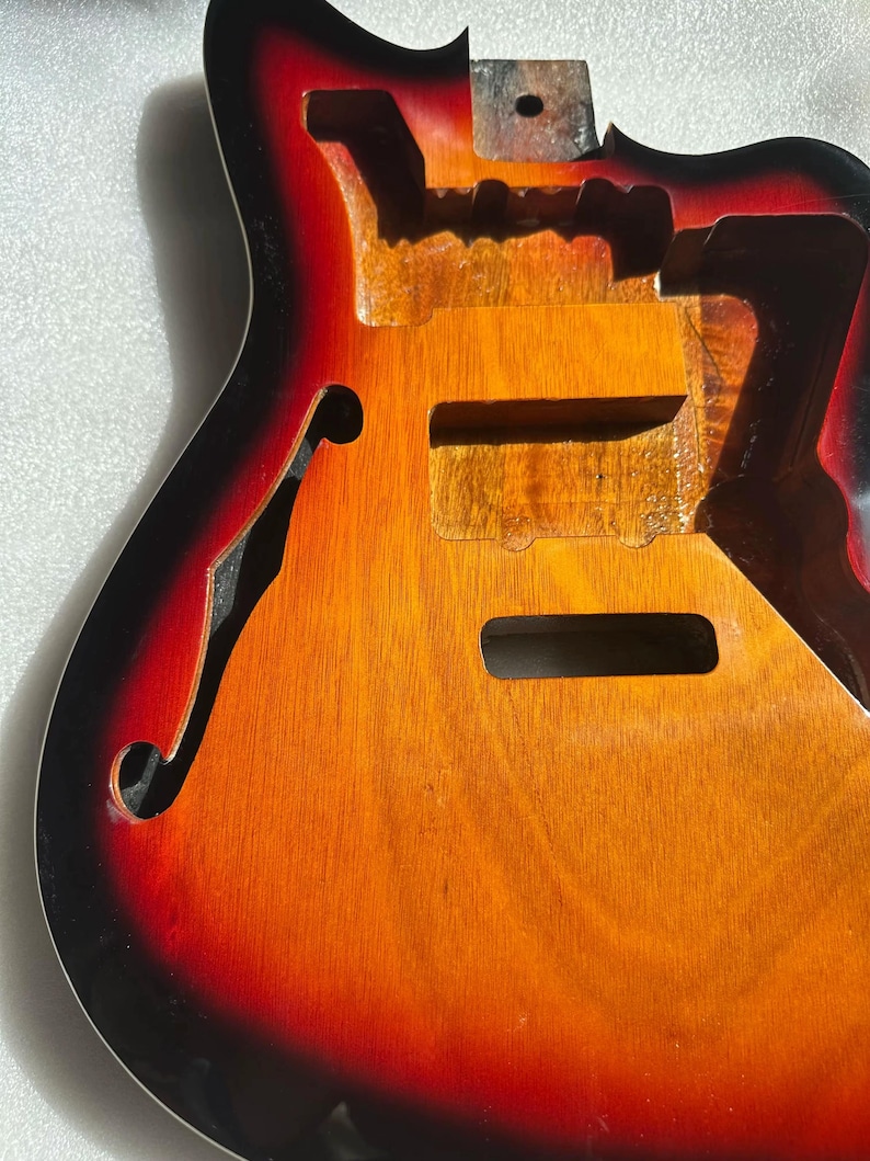 May include: A hollow-body electric guitar body with a sunburst finish. The body is unfinished, revealing the wood grain. The color transitions from black to red to orange and yellow. The guitar has a classic f-hole design and cutaways.