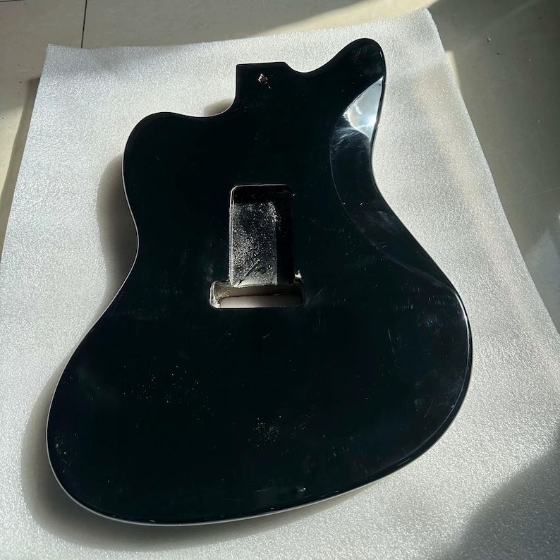 May include: A black guitar body with a white trim. The body has a cutaway design and a rectangular cavity for the electronics. The surface is glossy and reflects light. The guitar body is likely for a solid-body electric guitar.